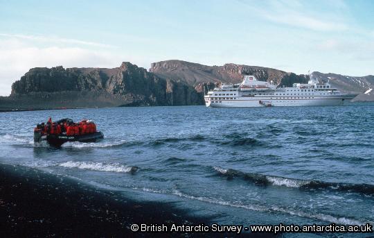 Tourists being taken ashore in an inflatable named Charles Darwin from tourist ship Hanseatic at Whalers bay, Deception Island , Cathedral Crags in background .