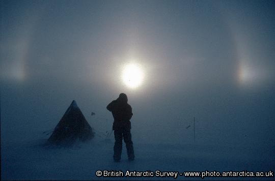 Parhelia over a camp site on the Evans Ice Stream In winds approaching gale force, ice crystals are picked up from the surface of the ice sheet and blown above head-height.  On this day the skies above the blowing snow were cloudless.  The light from the sun was refracted by the ice crystals to form parhelia, or sun-dogs. These winds heralded a week-long storm which this scientist sat out in his tent.
