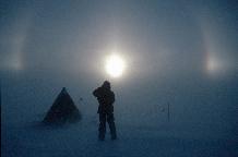 Parhelia over a camp site on the Evans Ice Stream In winds approaching gale force, ice crystals are picked up from the surface of the ice sheet and blown above head-height.  On this day the skies above the blowing snow were cloudless.  The light from the sun was refracted by the ice crystals to form parhelia, or sun-dogs. These winds heralded a week-long storm which this scientist sat out in his tent.